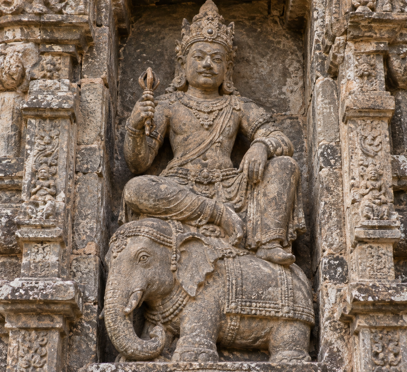 Close view of a crowned temple guardian holding a vajra with an elephant carved beneath him in the same stone niche.
