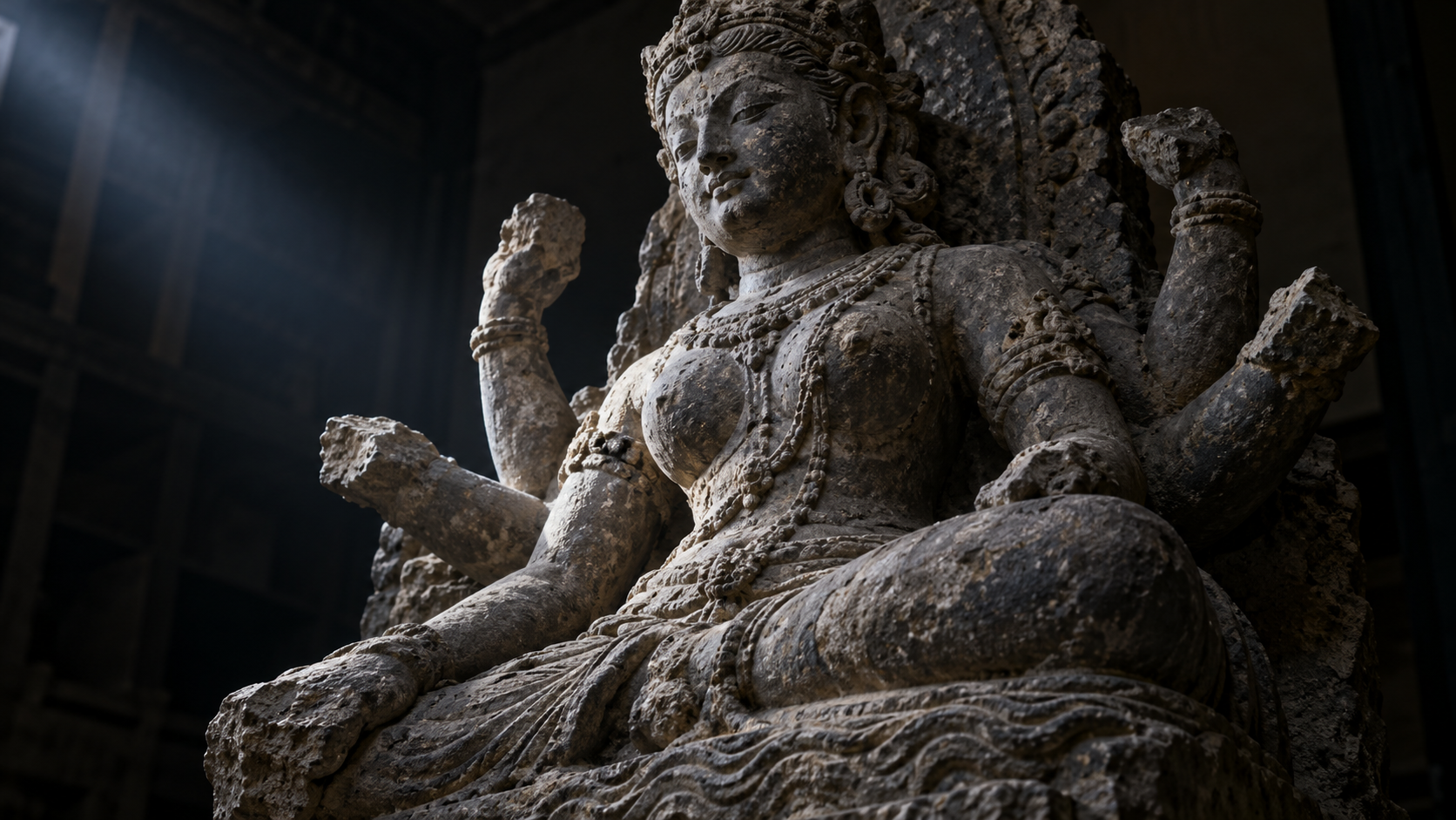 Close view of an eroded multi-armed goddess sculpture with broken hands and faint wave-like carving at its base.
