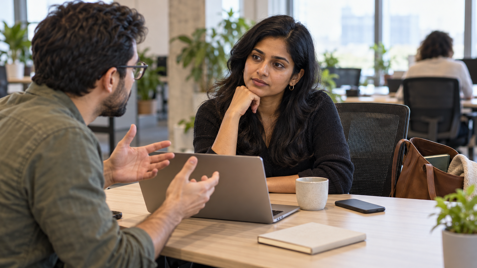 A professional pauses and listens calmly to a colleague during a difficult workplace conversation, with a closed book visible in a nearby bag.